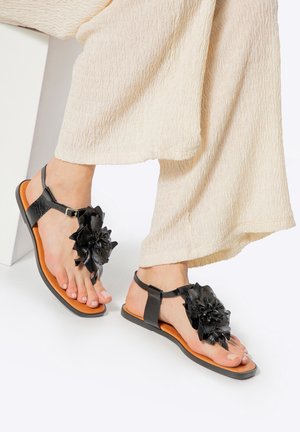 Voeten met zwarte sandalen met grote bloemdecoratie, gecombineerd met crèmekleurige, gestructureerde wijde pijpen broek op een witte achtergrond.