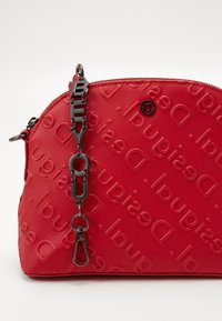 Sac rouge texturé avec logo en relief. Présente une chaîne noire avec le lettrage "SOIS TOI" , des accessoires en métal argenté et une fermeture à glissière arrondie.