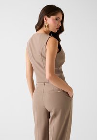 Beiger ärmelloser Overall mit tailliertem Oberteil und locker sitzender Hose. Verfügt über Gesäßtaschen und eine glatte Textur. Goldene Creolen als Akzente.