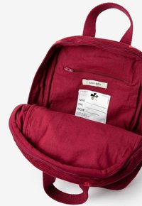 Intérieur d'un sac à dos en velours côtelé bordeaux avec une poche zippée, une étiquette pour les informations de contact et une poignée en textile. Texture lisse et forme arrondie.