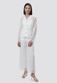 Conjunto de traje de encaje en blanco con mangas largas, botones en la parte delantera y dos bolsillos. Los pantalones tienen un ribete de encaje en el dobladillo. Combinado con tacones con tiras.