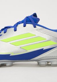 Futballcipők fényes ezüst felsőrésszel, kék kiegészítőkkel és neon zöld csíkokkal. Kék fűzőkkel és texturált mintázattal rendelkezik.