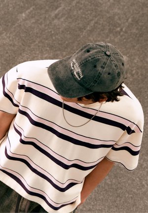 Person wearing white and purple striped shirt, gray cap with text, and chain necklace looking downward on a paved surface.