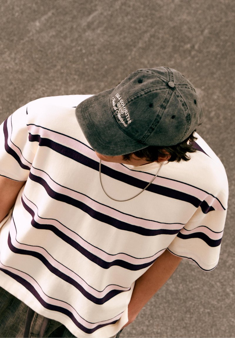 Personne portant une chemise rayée blanche et violette, une casquette grise avec un texte et un collier chaîne regardant vers le bas sur une surface pavée.