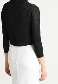 Suéter negro corto con mangas largas, de material texturizado, usado sobre una falda blanca con botones decorativos en la parte trasera.