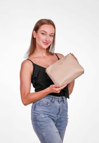 Krémová kožená kabelka na zip s vyraženým logem, kterou drží osoba v černém topu a modrých džínách, na bílém pozadí.