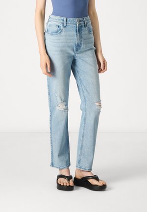 Lyse blå ripped jeans med høy midje og rette ben. Har fem lommer og knappelukking. Kombinert med svarte flip-flop sandaler.