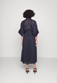 Nainen, jolla on kiharat hiukset, pukeutuneena löysään, tumman sävyiseen midi-mekkoon, jossa on leveät hihat, seisoo selin yksinkertaista taustaa vasten.