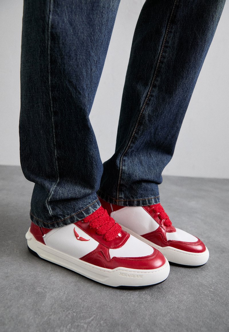 Red and white sneakers feature a chunky design, smooth leather material, textured laces, and branding on the sole. Worn with dark denim jeans.