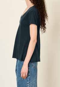 Žena s kudrnatými vlasy, oblečená v tmavozeleném tričku s krátkým rukávem a výstřihem do V, a modrých džínách, stojící bokem na neutrálním pozadí.
