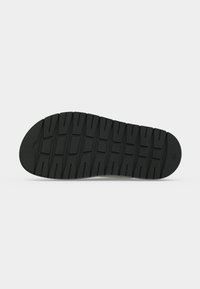 Semelle de chaussure noire avec des motifs de bande de roulement rectangulaires et un marqueur de taille rond près du talon, présentée sur un fond blanc uni.