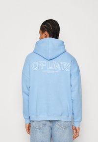 Felpa leggera blu con un design ampio e grande scritta stampata sul retro. Dotata di cappuccio con lacci e polsini a coste.