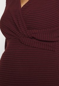 Gros plan d'une personne portant une robe portefeuille en maille côtelée rouge foncé avec un décolleté en V et un détail taille froncée.