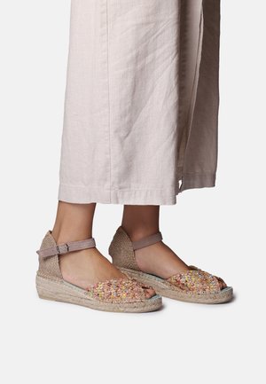 Piedi con sandali con zeppa in espadrille intrecciate multicolore con cinturini alla caviglia, abbinati a pantaloni larghi color beige chiaro.