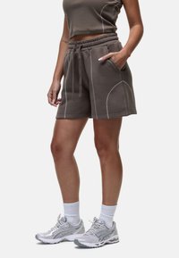 Shorts en coton marron avec un cordon de serrage à la taille et des poches latérales, avec des coutures blanches et une coupe décontractée. Accompagnés de chaussures de course grises.