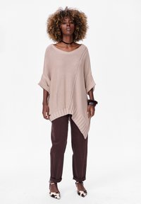 Beige oversized gebreide trui met een diagonale zoom en opgerolde mouwen, gecombineerd met donkerbruine broek en gebloemde hakken.