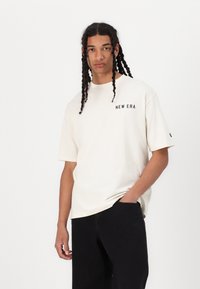 Cremefarvet oversized T-shirt med korte ærmer, som har et sort "NEW ERA" logo på venstre bryst, parret med sorte bukser.