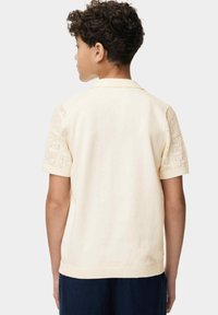Polo en un beige claro con mangas cortas y texturizadas. Presenta un cuerpo liso, un cuello y un dobladillo acanalado. Detalles intrincados en las mangas.