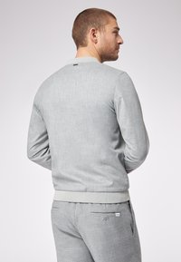 Felpa grigia lavorata a maglia con una texture liscia, polsini e orlo a coste, e un piccolo logo sulla parte posteriore del collo. Indossata con pantaloni grigi a fantasia.