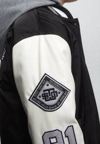 Czarno-biała kurtka bomberka z szarym kapturem. Posiada naszywkę na rękawie z tekstem i wyraźną cyfrą. Gładka faktura materiału.