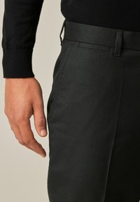 Sorte mønstrede bukser med en tekstureret overflade; et nærbillede viser taljen og en hånd, der hviler på benet.