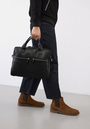 Henkilö, jolla on musta takki ja tummat farkut, pitää mustaa nahkaista salkkua ja jalassa on ruskeat mokkasaappaat vaalealla lattialla.