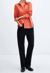 Camicia coral a maniche lunghe con bottoni, abbinata a jeans neri larghi e scarpe nere, che mettono in risalto un fit rilassato e una texture morbida.