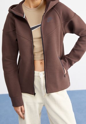 Personne portant un sweat à capuche marron zippé sur un t-shirt Nike beige coupé, et un pantalon en velours côtelé crème, debout avec une main dans la poche.