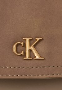 Χρυσό μεταλλικό λογότυπο "cK" σε ανοιχτό καφέ δερμάτινη επιφάνεια με ορατά ραψίματα κοντά στην κάτω άκρη.