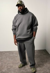 Grijze sweatshirt met capuchon, over een zwart shirt gedragen. Bijpassende grijze broek, groene sneakers en een groene pet met een wit logo.