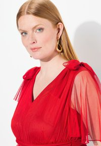 Červené šaty s průhlednými, volánkovými rukávy a hlubokým výstřihem do V. Hladká tkanina, pásek v pase, doplněné velkými zlatými náušnicemi.