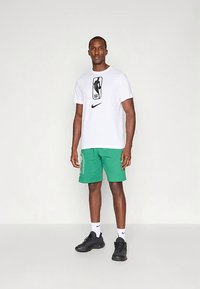 Fehér Nike póló NBA logóval, zöld rövidnadrággal, oldalán felirattal. Az outfitt fekete sportcipő és fehér bokazokni teszi teljessé.