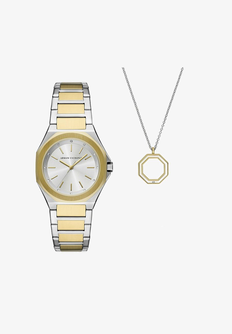 Stříbrně a zlatě tónovaný osmiúhelníkový hodinky s nerezovým páskem, s stříbrným ciferníkem a zlatým akcentem na lunetě. Součástí je zlatý osmiúhelníkový přívěsek na stříbrném řetízku.