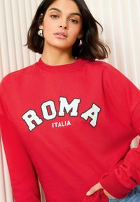Il felpa rossa presenta un coltondo e la scritta "ROMA ITALIA" ricamata in bianco e nero. Il tessuto è morbido e ha una vestibilità relax.