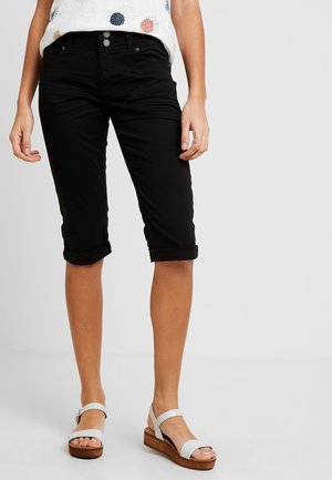 Femme portant un short noir retroussé jusqu'aux genoux, un haut blanc à motifs et des sandales à plateforme blanches avec des semelles en bois.