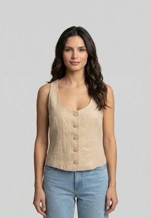 Mujer de cabello largo y oscuro, que lleva un top sin mangas beige con botones y jeans azul claro, de pie frente a un fondo gris liso.