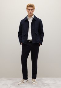 Giacca in misto lana di colore navy con chiusura a bottone e due tasche frontali, indossata sopra una camicia bianca e pantaloni scuri, completata da sneakers beige.