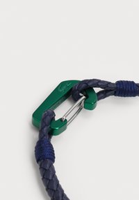 Bracciale in pelle intrecciata blu navy con chiusura in metallo verde dotata di logo e dettagli in hardware argentato. La struttura è flessibile ma resistente.