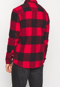 Camicia in flanella a quadri rossi e neri con colletto a bottoni, maniche lunghe e orlo arrotondato. Materiale in cotone con una consistenza morbida.