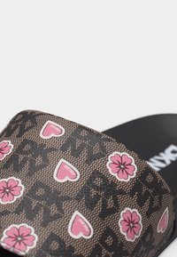 Gros plan sur la lanière d'une sandale noire à enfiler ornée de logos DKNY, de cœurs roses et de fleurs roses imprimés sur un fond marron texturé.