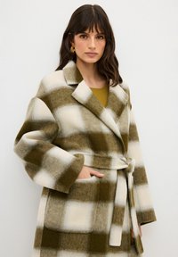 Cappotto in misto lana a quadri con rever oversize, vita con cintura e tasche. La combinazione di colori include verde oliva e crema con una texture morbida.