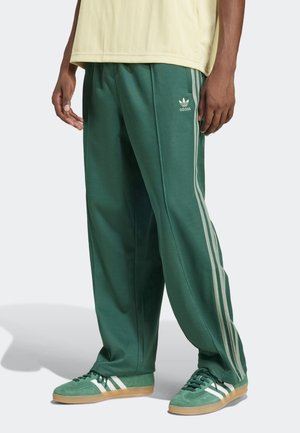 Person wearing green Adidas track pants with white stripes and matching green sneakers with white detailing on a plain background.