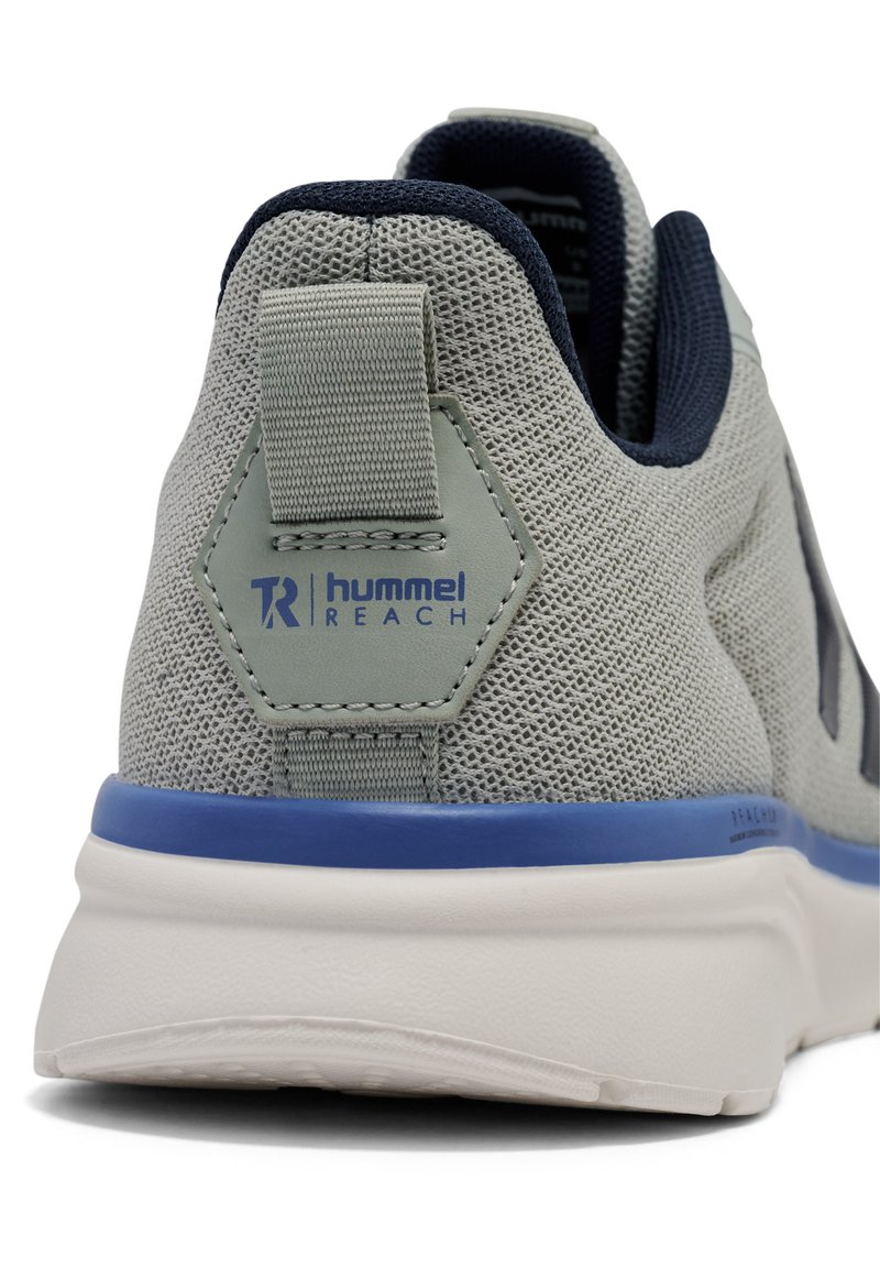 Zapatilla de malla gris claro con una pestaña en el talón en forma hexagonal con acentos azules y logo, suela de goma contrastante y detalles en azul marino en el acolchado del tobillo.