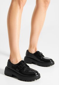 Zapatos de charol negro con una suela gruesa y acanalada y un detalle de hebilla en la parte superior. Textura suave y forma de punta redondeada.