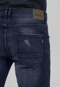 Dark blue denim jeans with a faded texture, a patch on the back waist detailing the brand "Blend," and a sculpted back pocket.