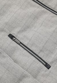 Szara tkanina w kratę z czarną kieszenią na suwak i metalowymi elementami. Tekstura jest miękka, a wzór geometryczny z subtelnymi odcieniami.