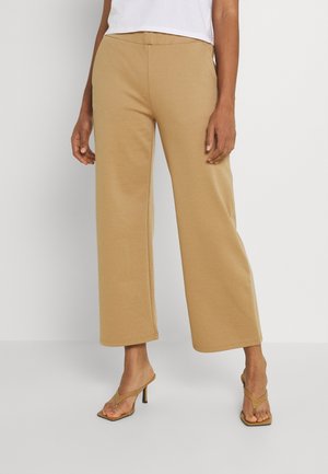 Kvinde iført lysebrune bukser med vide ben og matchende høje hæle, står mod en hvid baggrund.