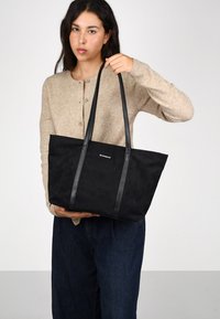 Bolso de mano negro con textura de ante suave, que cuenta con dos asas de cuero, diseño minimalista y un logo sutil en la parte frontal.