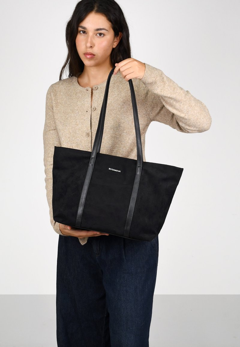 Bolso de mano negro con textura de ante suave, que cuenta con dos asas de cuero, diseño minimalista y un logo sutil en la parte frontal.