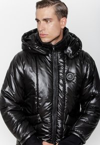 Mężczyzna w błyszczącej czarnej kurtce puchowej z kapturem, zapiętej, patrzący w bok na tle jednolitego białego tła.